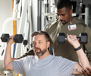 Man coaching another man doing strength-training exercise with hand weights.