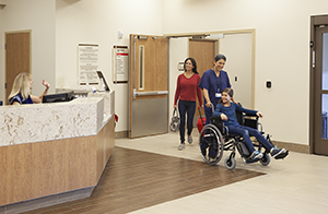 Health care provider pushing girl in wheelchair toward hospital exit with woman walking alongside.