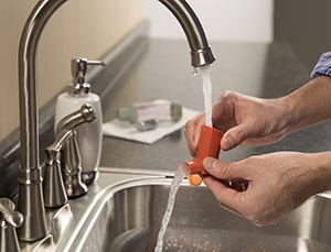 Hand holding inhaler mouthpiece under running water. Spacer lying on towel on counter.