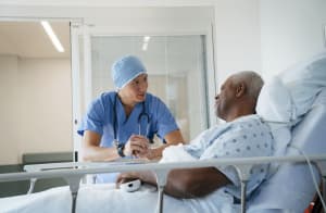 In a hospital, the patient wears a hospital gown and rests in a hospital bed while talking with the doctor in scrubs.